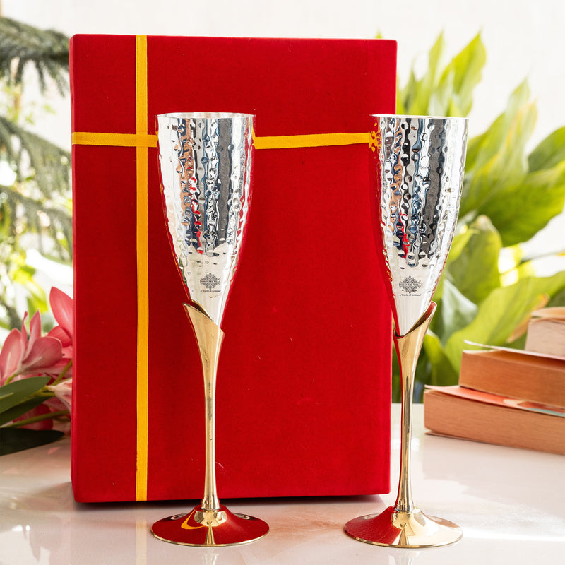 Two silver champagne flutes with gold bases in front of a red gift box on a white surface with greenery in the background.