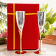 Two silver champagne flutes with gold bases in front of a red gift box on a white surface with greenery in the background.